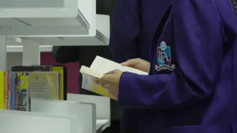 BBC A student holds a book open in a school library.