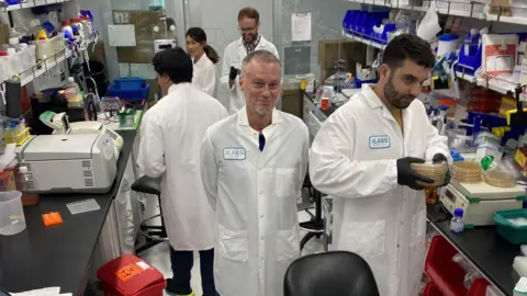 Jacob Glanville Tim Friede in a lab coat stands in the middle of a busy laboratory where four other researchers are working